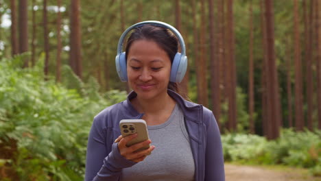 Woman-Wearing-Wireless-Headphones-Exercising-Doing-Work-Out-Outdoors-Streaming-Music-Or-Podcast-From-Mobile-Phone-Running-Along-Track-In-Forest-Wearing-Sports-Clothing-Shot-In-Real-Time